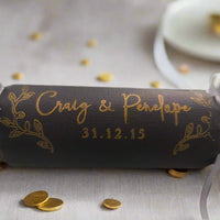 Black wedding cracker with gold foil personalisation, featuring names and date, tied with silver ribbons, styled with metallic gold leaves on a grey background.