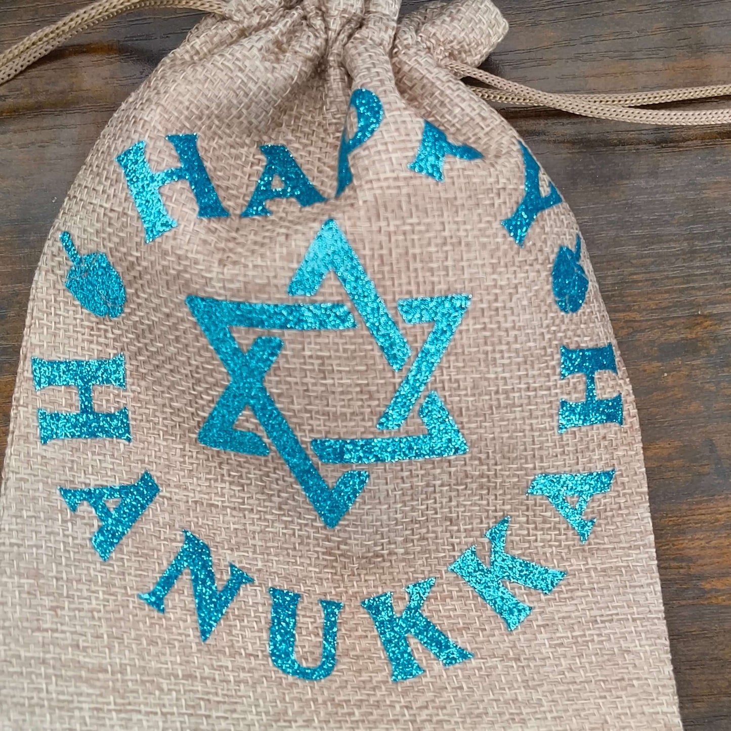 Side angle of cinched Hanukkah gift bag showing blue glitter Happy Hanukkah design on hessian fabric