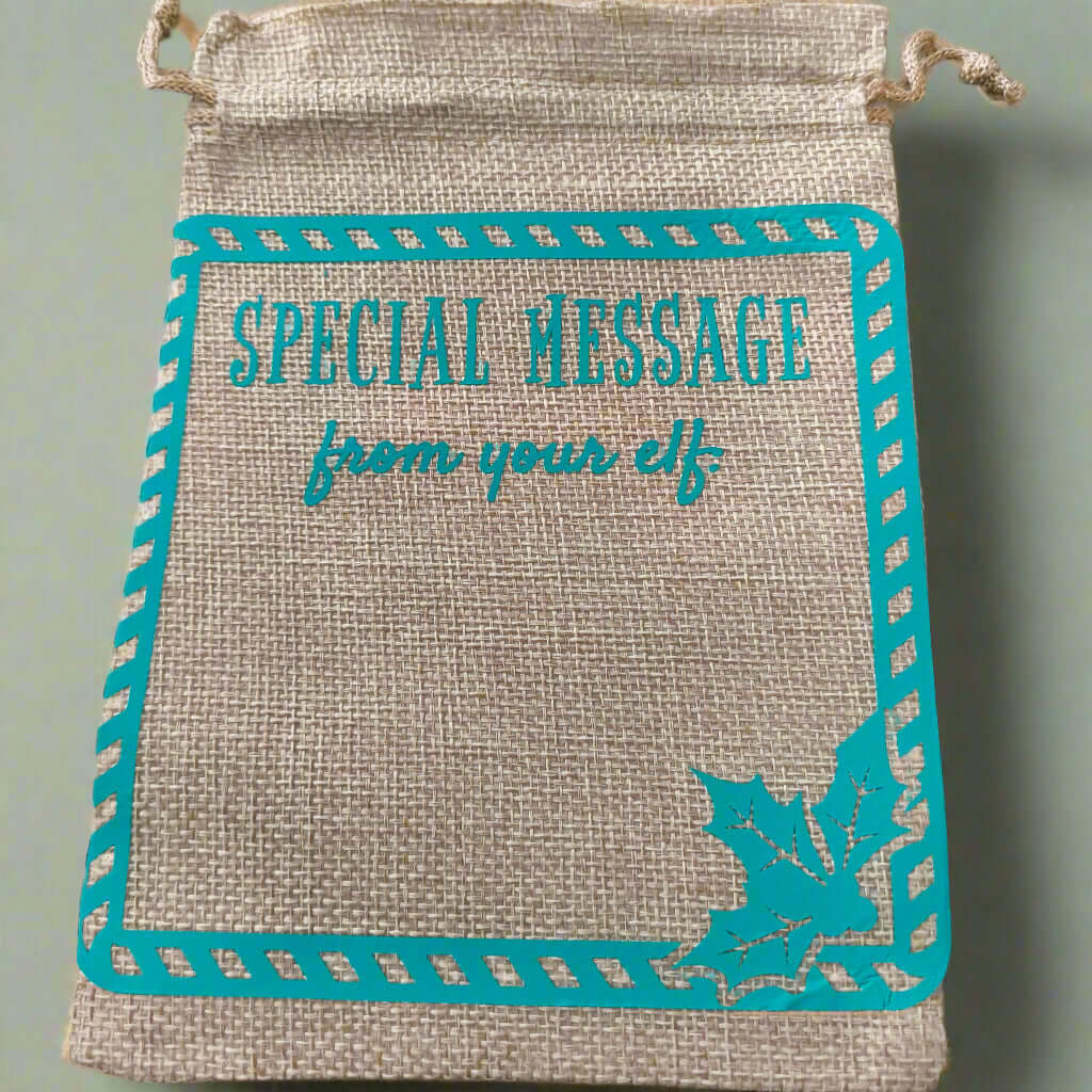 Burlap drawstring gift bag with turquoise vinyl text reading 'Special Message from your elf', framed with a candy cane-style border and a holly leaf design in the bottom right corner.