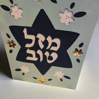 A light blue card stands upright, featuring a dark blue Star of David cut out in the center. The Hebrew text "מזל טוב" (Mazel Tov) is prominently displayed within the star in white. The card is decorated with cut-out flowers in white, dark blue, and metallic brown.