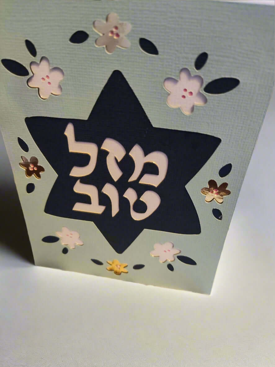 A light blue card stands upright, featuring a dark blue Star of David cut out in the center. The Hebrew text "מזל טוב" (Mazel Tov) is prominently displayed within the star in white. The card is decorated with cut-out flowers in white, dark blue, and metallic brown.