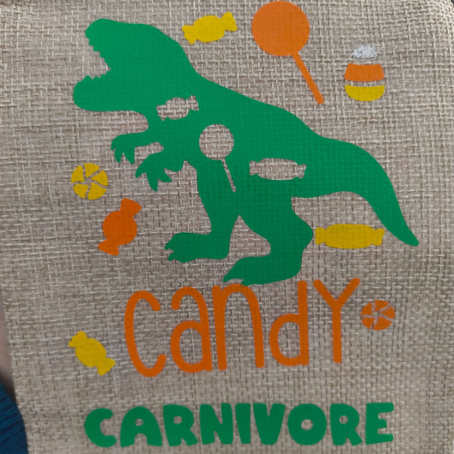 Close-up of a hessian fabric design featuring a green dinosaur silhouette surrounded by colourful sweets and the words 'Candy Carnivore' in orange and green lettering