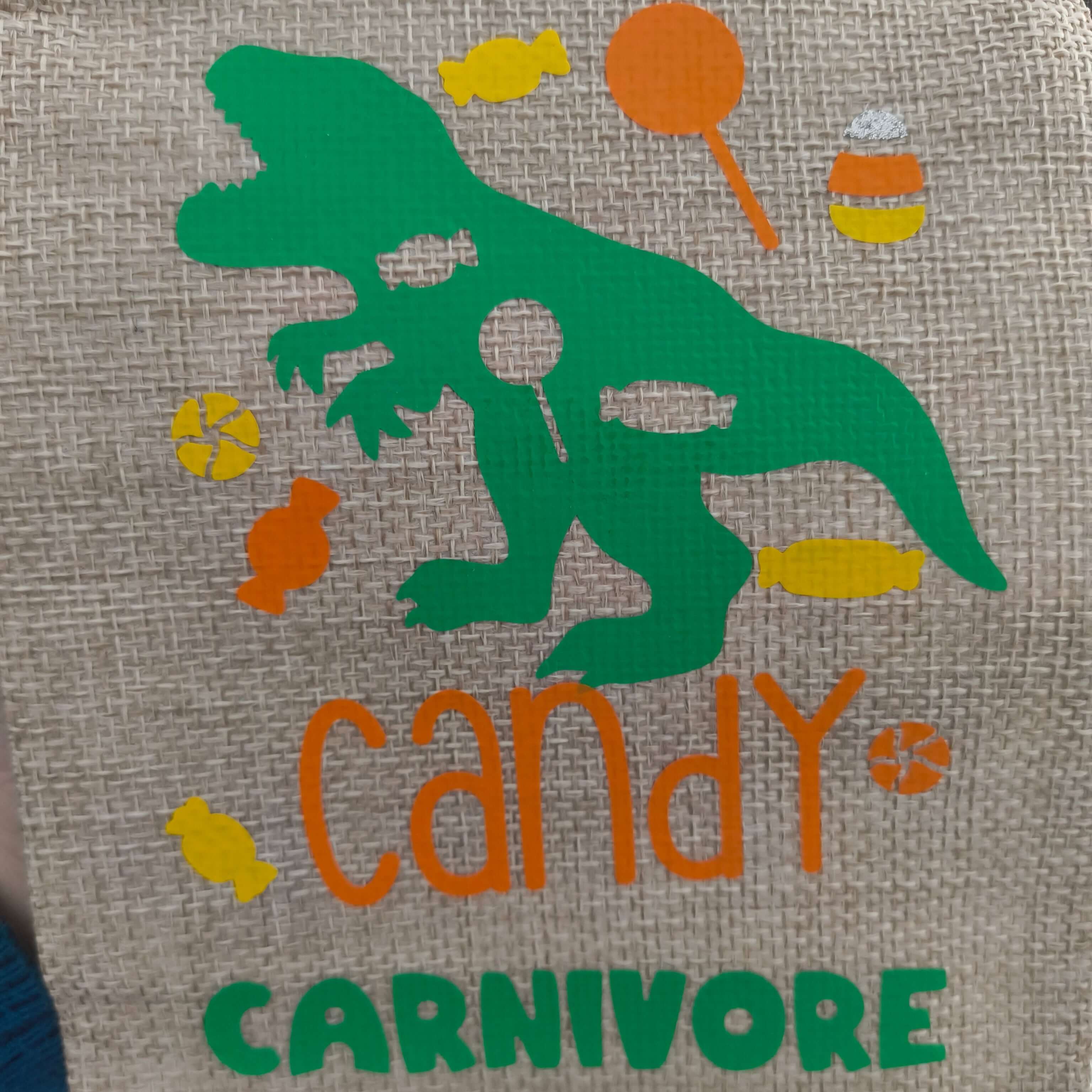 Close-up of a hessian fabric design featuring a green dinosaur silhouette surrounded by colourful sweets and the words 'Candy Carnivore' in orange and green lettering