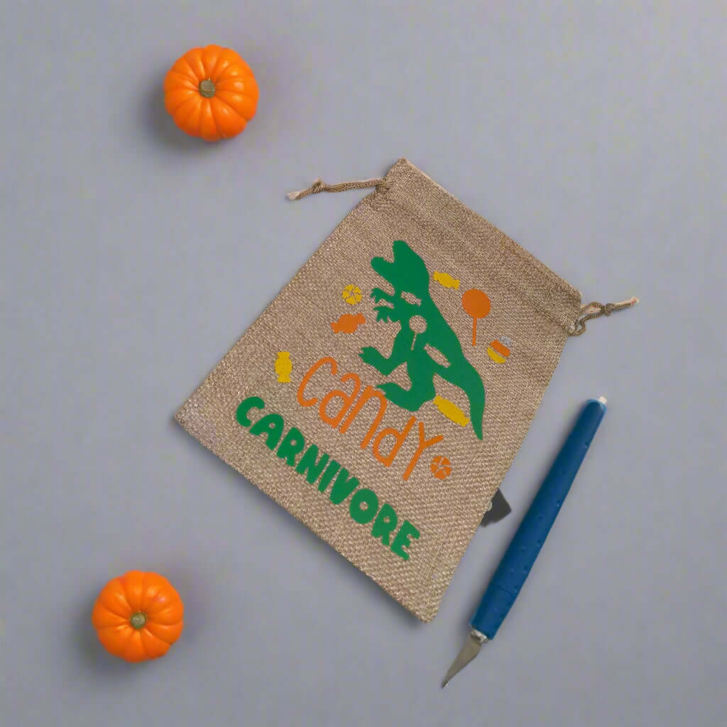 "Handmade hessian drawstring bag with a green dinosaur design and the phrase 'Candy Carnivore' in orange and green lettering. The bag is placed on a grey background with two small orange pumpkins and a craft knife beside it.