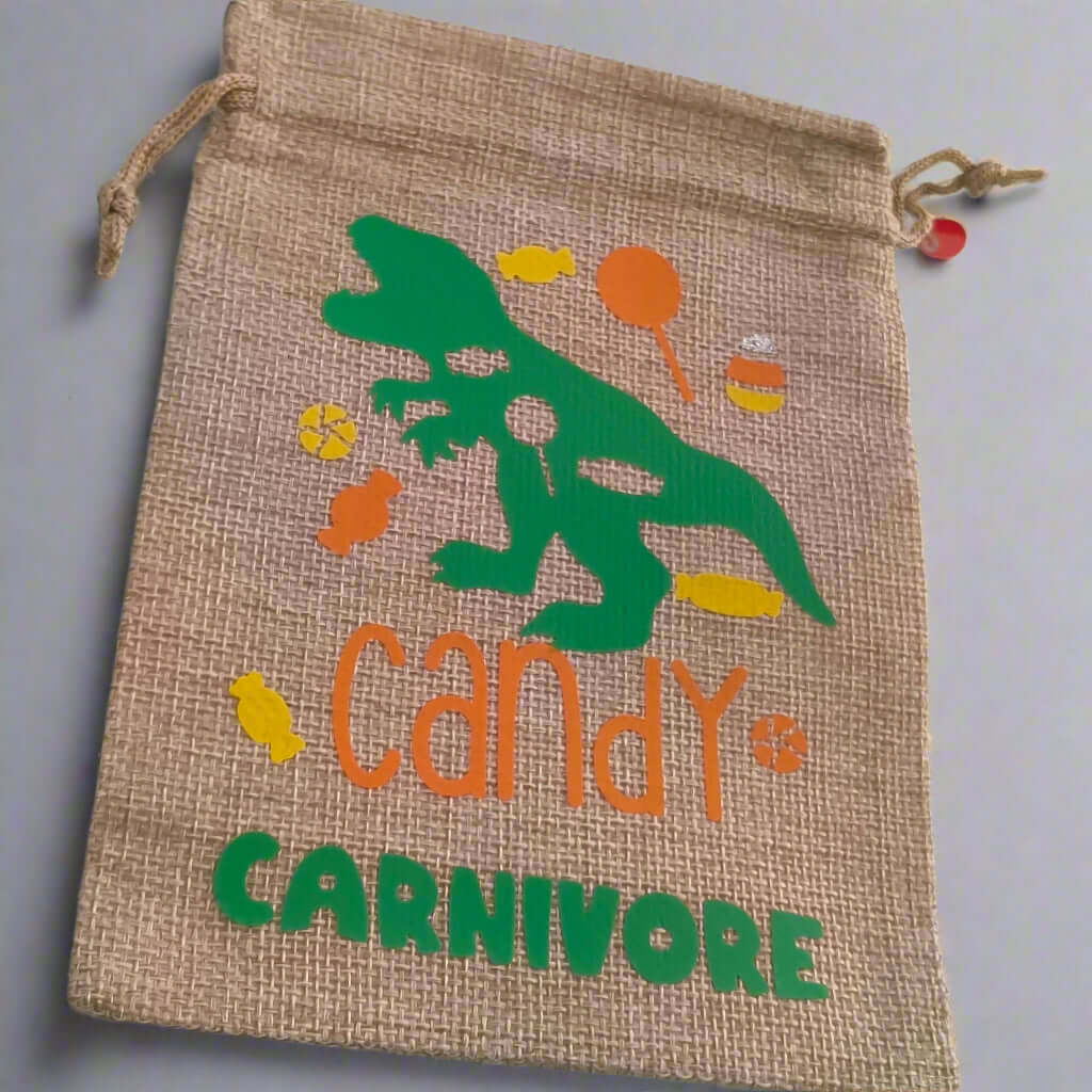 Handmade hessian drawstring bag with a green dinosaur graphic surrounded by colourful sweets and the phrase 'Candy Carnivore' in orange and green text. The bag is displayed on a plain light grey background.
