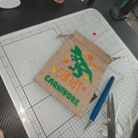 Handmade hessian drawstring bag with a green dinosaur and the words 'Candy Carnivore' in orange and green vinyl. The bag is displayed on a craft cutting mat, surrounded by crafting tools including a precision knife, scissors, and vinyl scraps.