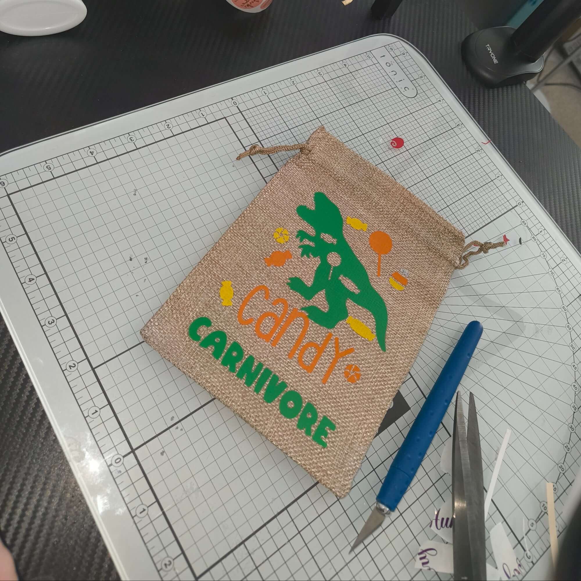 Handmade hessian drawstring bag with a green dinosaur and the words 'Candy Carnivore' in orange and green vinyl. The bag is displayed on a craft cutting mat, surrounded by crafting tools including a precision knife, scissors, and vinyl scraps.