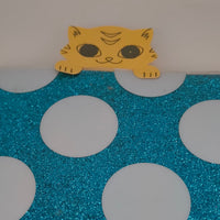 Handmade yellow cat paper cut-out peeking over the edge of a blue glittery polka dot surface, resembling a bookmark.
