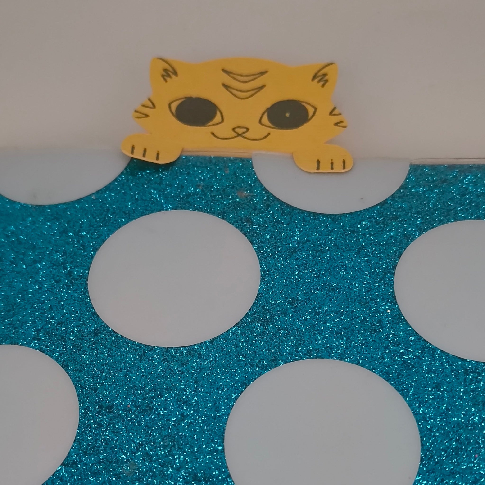 Handmade yellow cat paper cut-out peeking over the edge of a blue glittery polka dot surface, resembling a bookmark.
