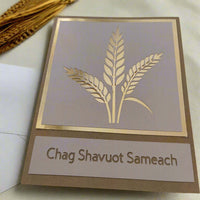 Handmade "Chag Shavuot Sameach" greeting card with a metallic gold wheat design, displayed on a light fabric surface with a small bundle of wheat stalks and an envelope in the background.