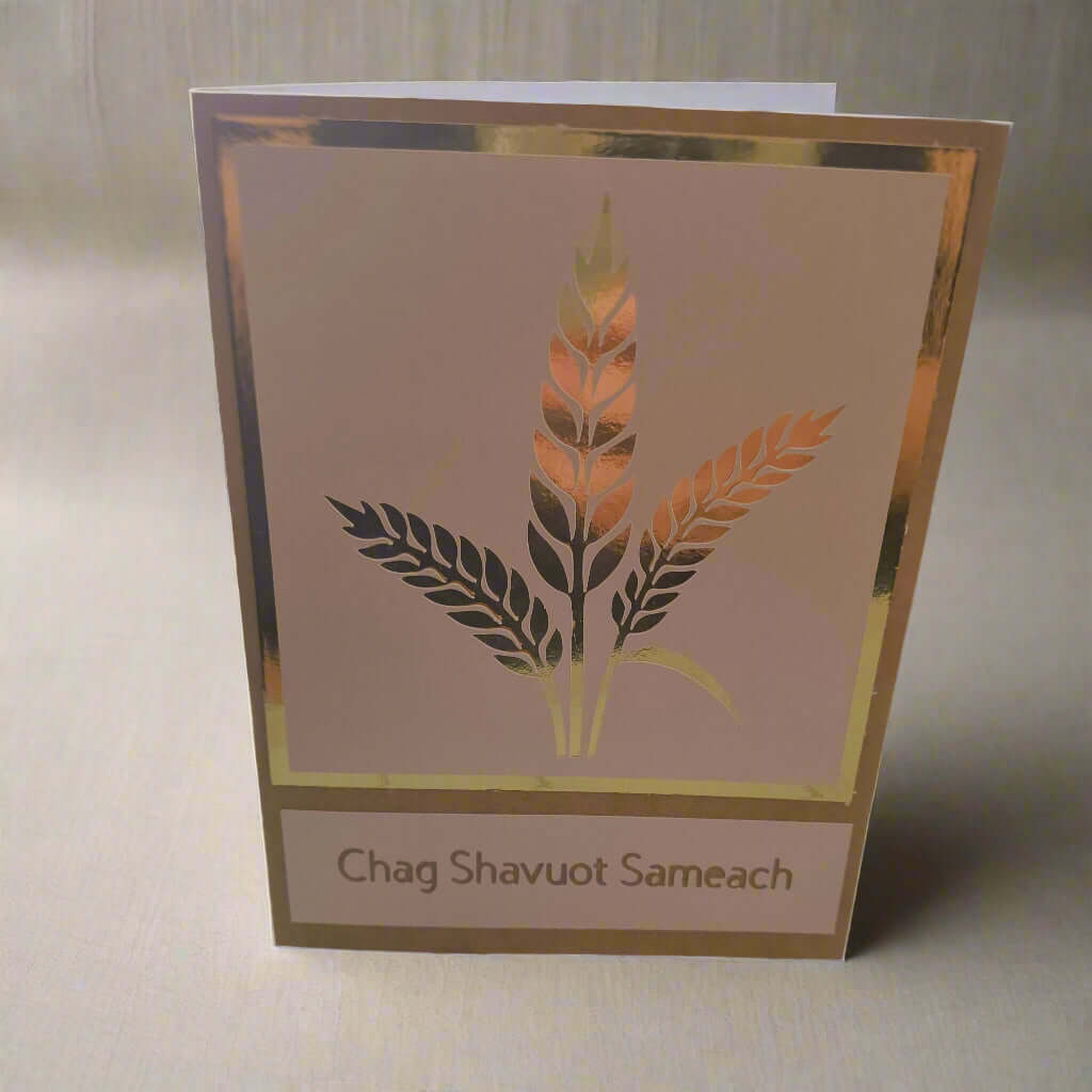 A handmade "Chag Shavuot Sameach" greeting card features a gold and copper metallic wheat design bordered by a gold foil frame on a beige background. The card stands upright on a soft, textured, light beige fabric surface.