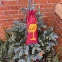 Cheers wine gift bag in red hessian with gold glitter design shown outdoors in garden setting.