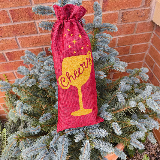 Red hessian wine bag with gold glitter "Cheers" design displayed on a pine tree.