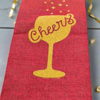 Red wine bottle gift bag with gold glitter 'Cheers' design, photographed from a top-down angle on a grey background with curling gold ribbon for decoration.