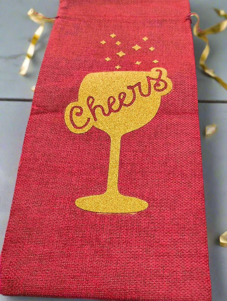 Red wine bottle gift bag with gold glitter 'Cheers' design, photographed from a top-down angle on a grey background with curling gold ribbon for decoration.