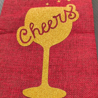 Close-up of a red hessian wine bottle bag featuring a gold glitter 'Cheers' wine glass design, viewed from a front-on angle, showcasing the texture and sparkle detail.

