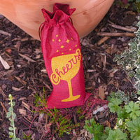 Red hessian cheers wine bottle bag with gold glitter design placed in garden among greenery.