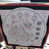 Chinese New Year greeting card with dragon design, lanterns, and cherry blossoms, layered red and black border, angled front view on lace tablecloth