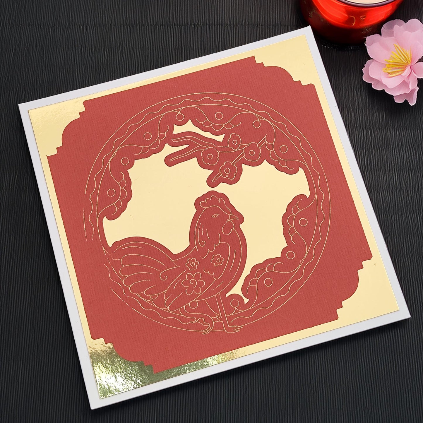 Angled top-down view of a red and gold Chinese New Year rooster card on a black placemat, handmade with layered cardstock, ideal for Lunar New Year greetings.