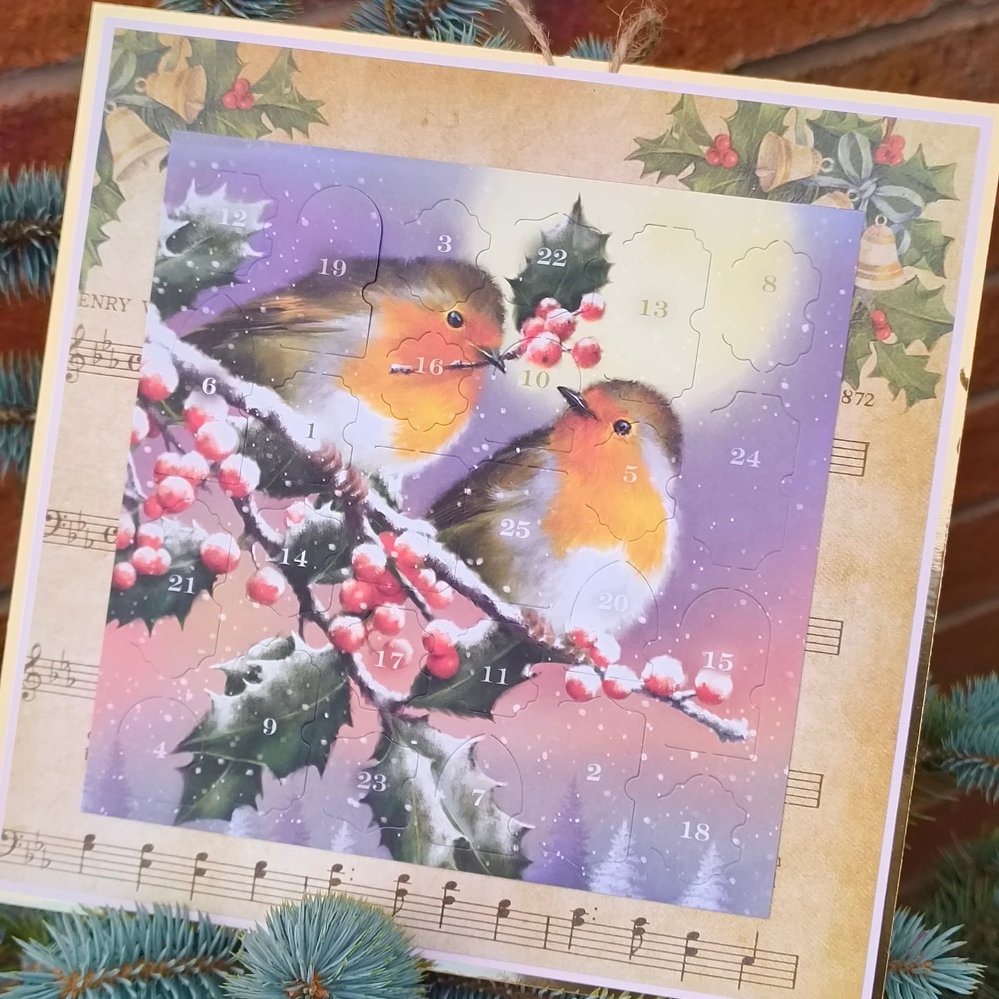 Handmade Christmas advent calendar featuring two robins perched on a snowy holly branch with red berries, displayed against a brick wall and evergreen foliage.