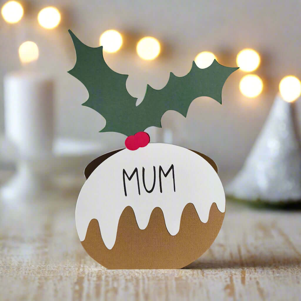 Christmas pudding-shaped place card with “Mum” written on it, decorated with green holly leaves and red berries, standing on a rustic wooden table with festive lights in the background.