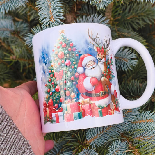 White ceramic mug with festive Christmas scene featuring Santa, reindeer, decorated tree, and presents, hand holding mug