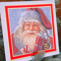 Classic Santa Claus Advent Calendar with 25 numbered doors, red and gold border, resting on a pine tree branch with natural outdoor lighting.