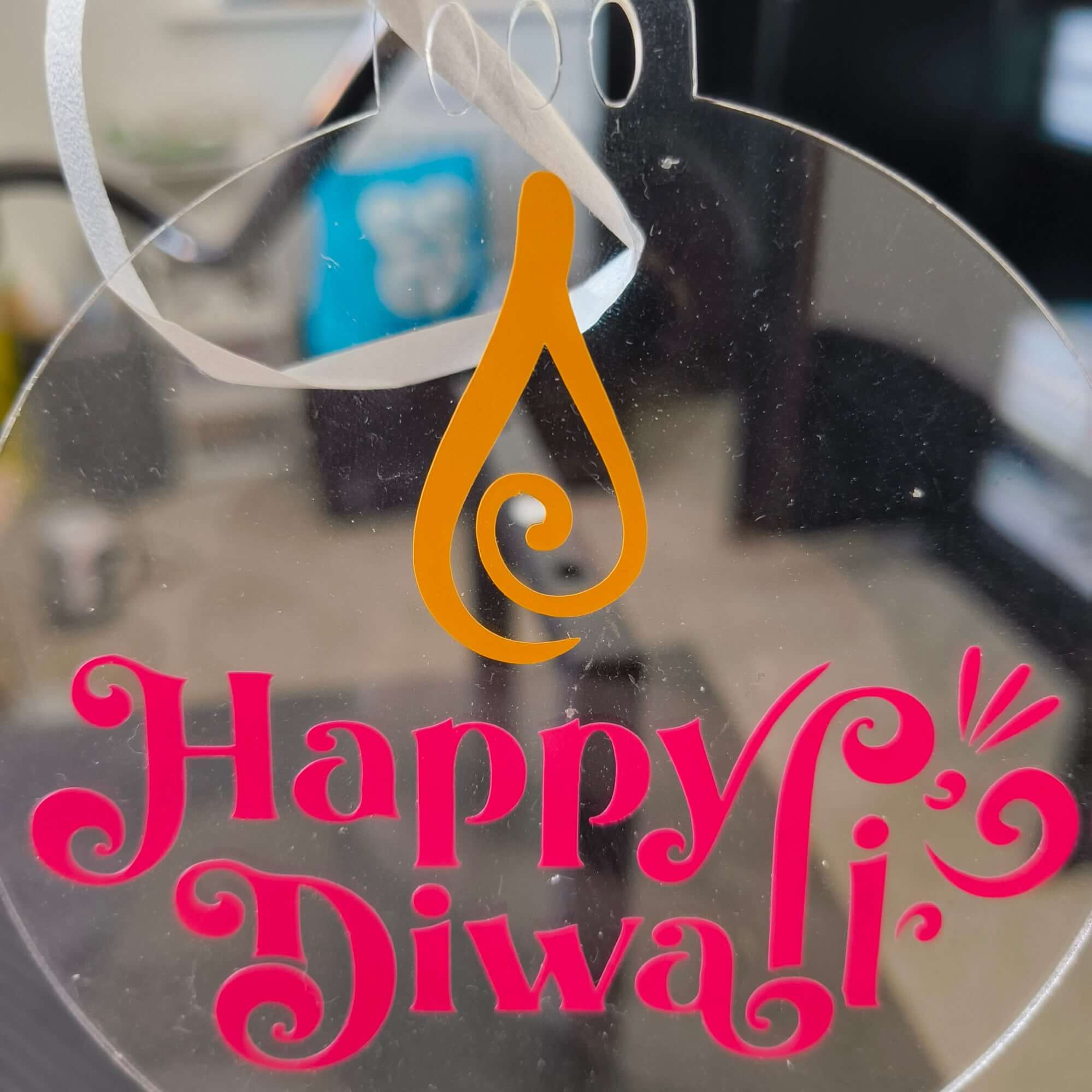 Close-up of clear acrylic Happy Diwali bauble with pink text and orange diya flame design, ideal for Hindu festive decor and gifts.