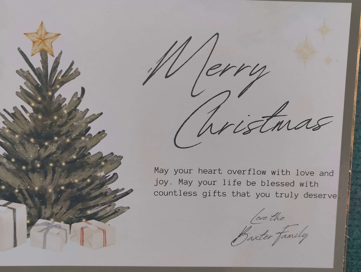 Close-up of a luxury handmade Christmas card featuring a decorated tree with a gold star topper and glowing lights, surrounded by wrapped presents. The card bears the message “Merry Christmas” in elegant script, with a heartfelt sentiment printed below and signed “Love the Baxter Family.” The design is bordered with gold and green details.