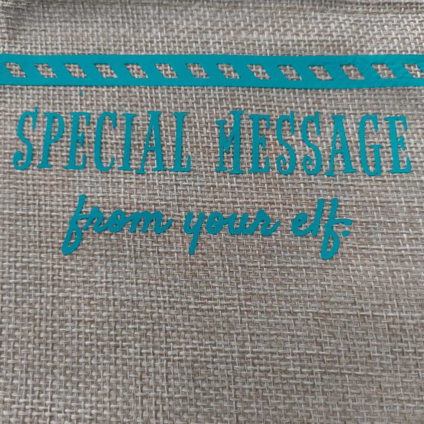 Close-up of turquoise vinyl text on a burlap surface reading 'Special Message from your elf', with part of a striped decorative border visible at the top