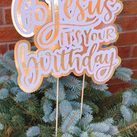 Close-up of a gold and white “Go Jesus It’s Your Birthday” cake topper with script lettering on a spruce tree.