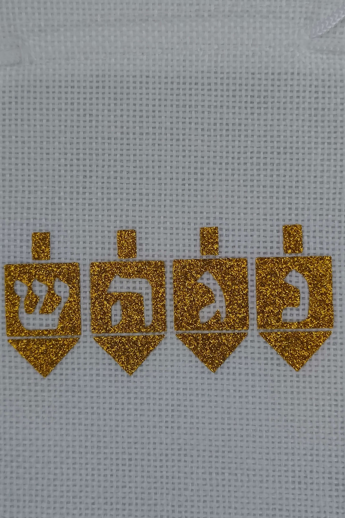 Close-up view of four golden glitter dreidels with Hebrew letters (Shin, Hey, Gimel, Nun) ironed onto white textured hessian fabric. The sparkling vinyl contrasts against the crisp weave of the bag, celebrating the Hanukkah tradition with a handmade feel.