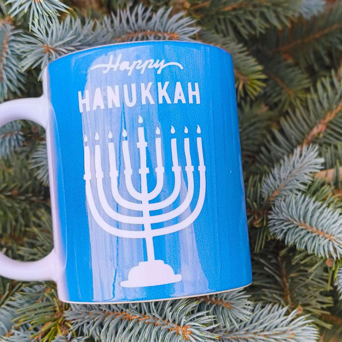 Happy Hanukkah mug featuring a menorah design on a blue background among evergreen branches.