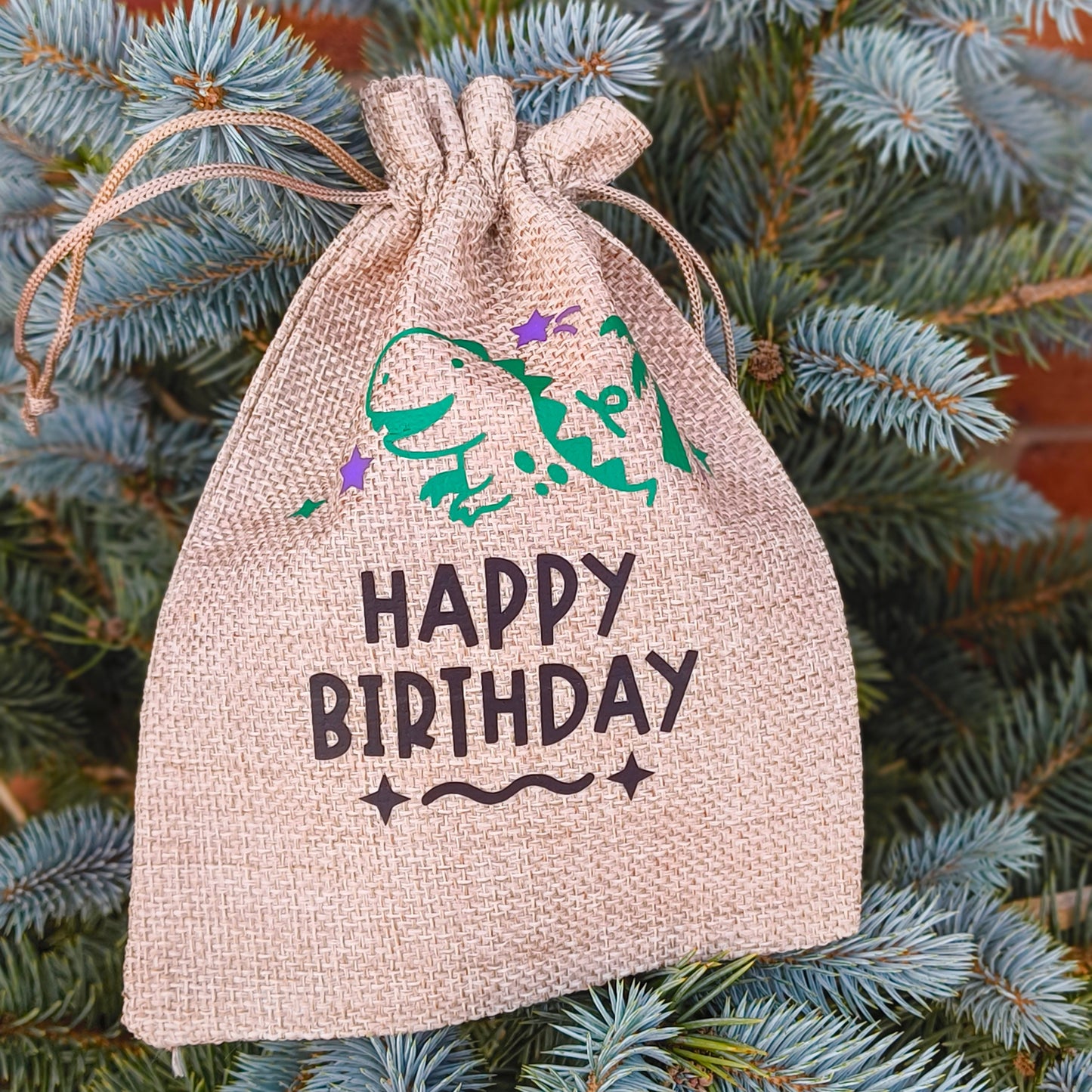 Close-up of hessian Happy Birthday gift bag with green dinosaur and purple stars, rustic design on blue spruce tree.