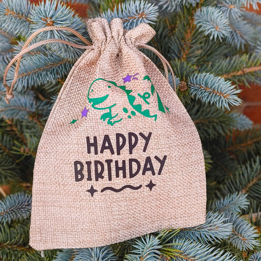 Close-up of hessian Happy Birthday gift bag with green dinosaur and purple stars, rustic design on blue spruce tree.