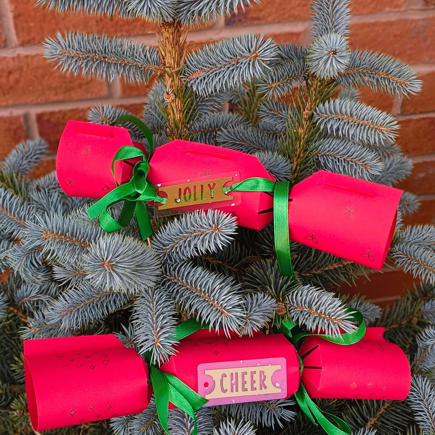 Close-up of red Christmas crackers tied with green ribbon and gold Jolly and Cheer tags, displayed on a blue spruce tree