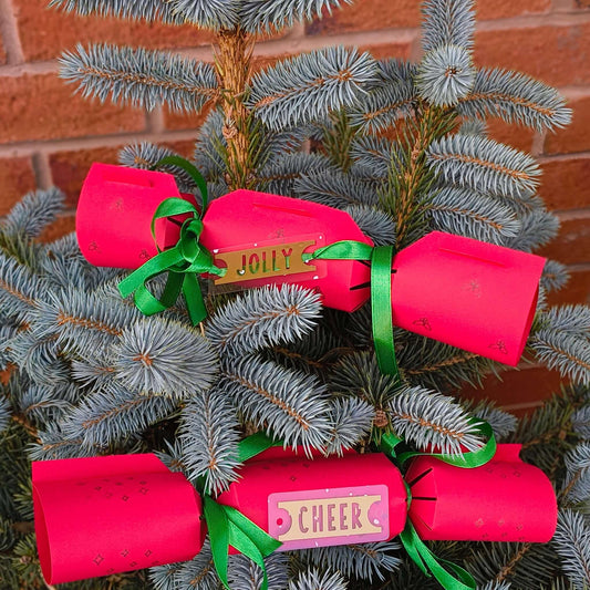 Close-up of red Christmas crackers tied with green ribbon and gold Jolly and Cheer tags, displayed on a blue spruce tree