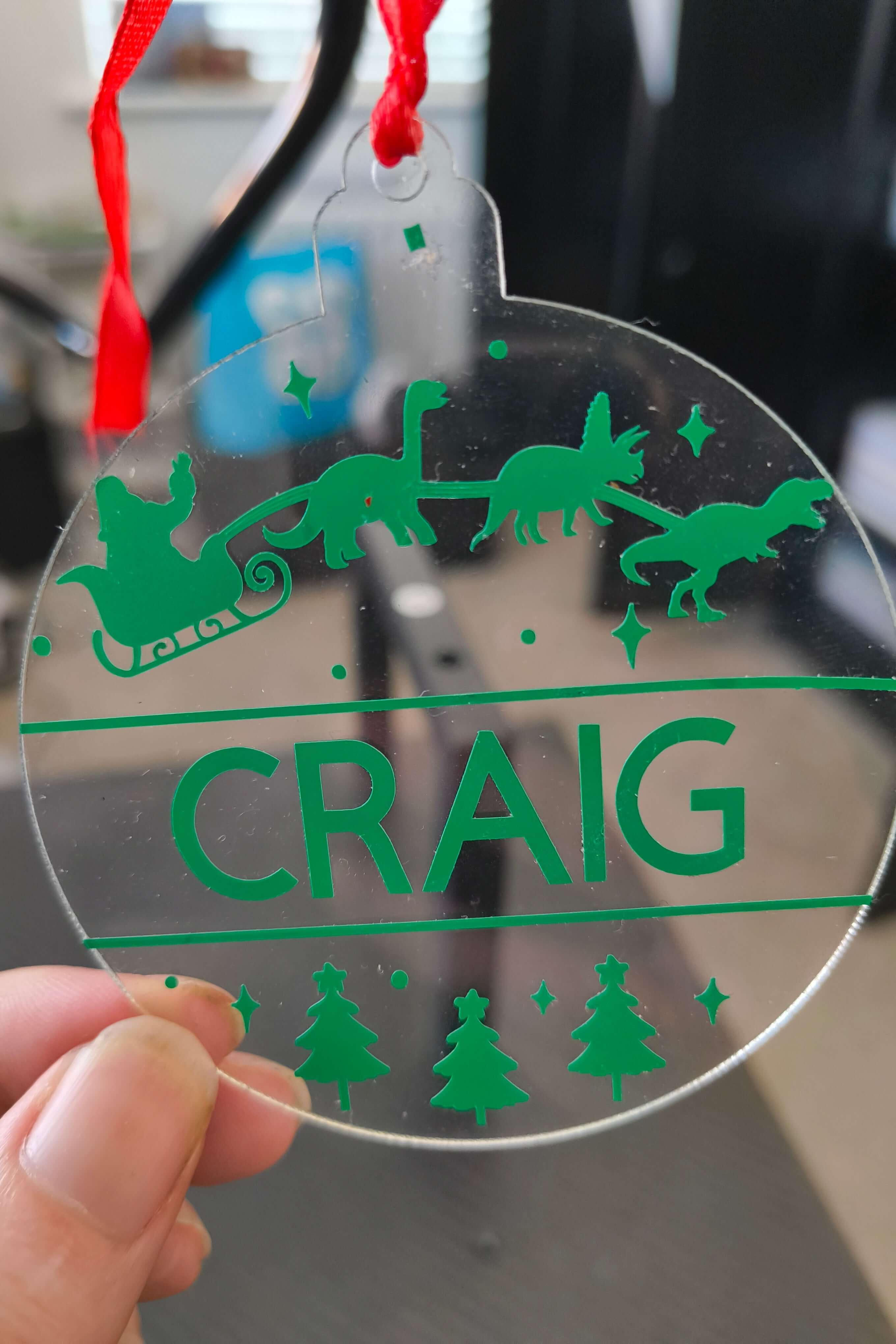 Close-up of personalised acrylic dinosaur Christmas bauble in green vinyl, featuring Santa's sleigh and festive trees.