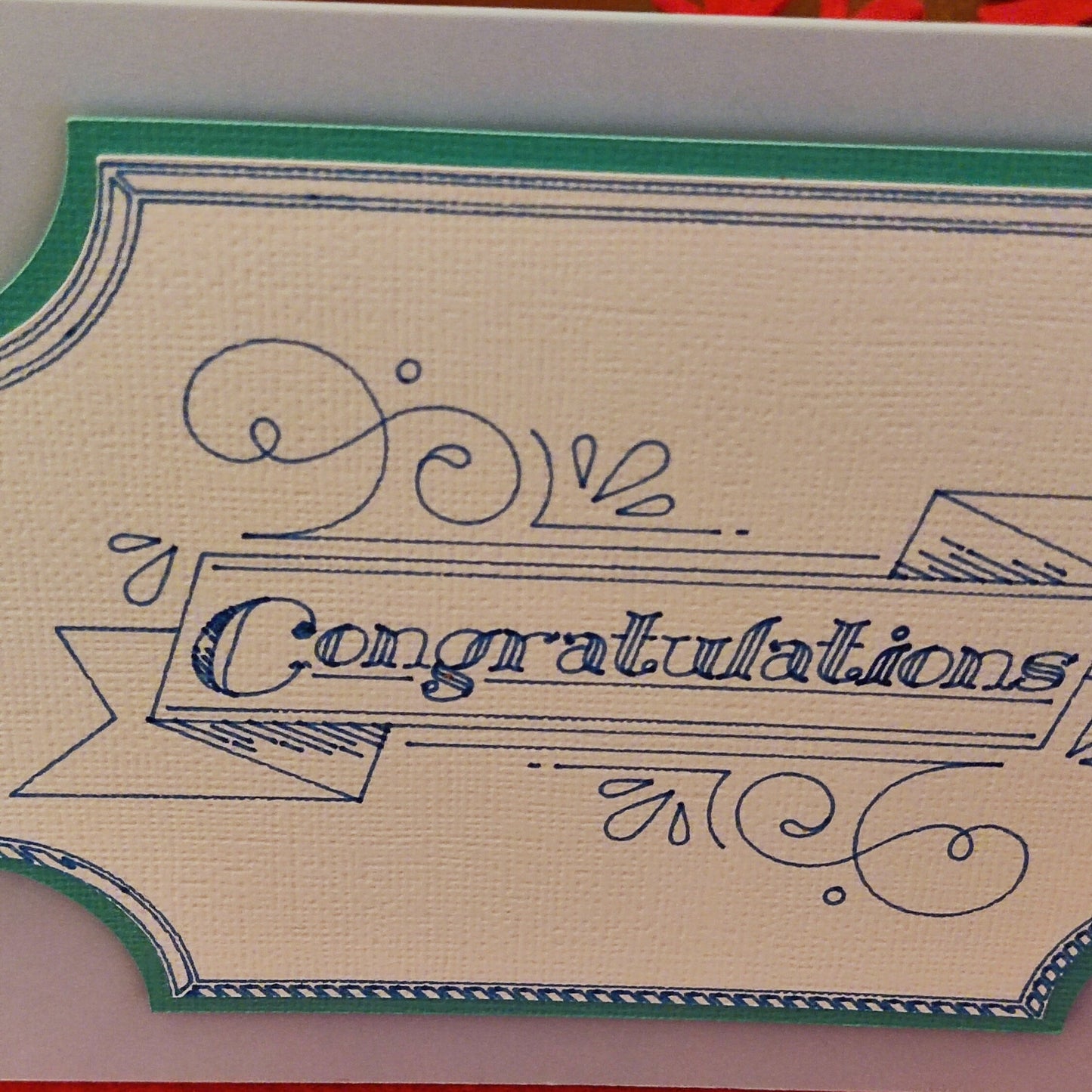 Close-up of the ornate banner design with “Congratulations” text, printed in blue on textured ivory card with a teal border. Highlights the intricate detail of the handmade finish.