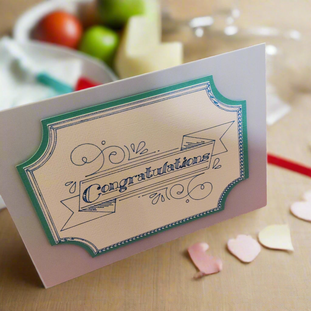 Greeting card featuring “Congratulations” in a decorative blue script with teal edging. Photographed beside fruit and craft supplies for scale, showing its medium size.