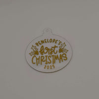 Personalised first Christmas bauble in white acrylic with gold text and holly design on plain background.
