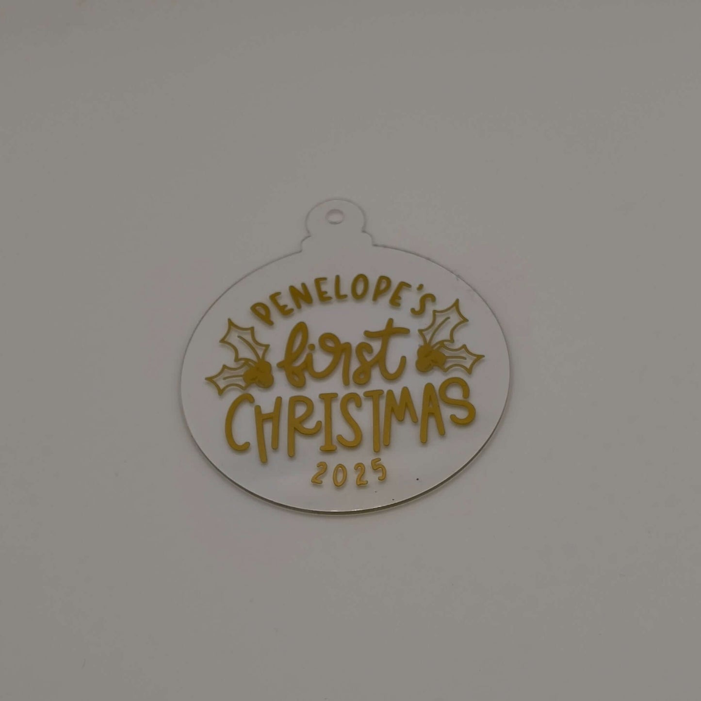 Personalised first Christmas bauble in white acrylic with gold text and holly design on plain background.
