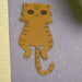 Top-down view of a cute orange tiger cat paper bookmark with black stripes, placed on a grey background beside a book and green leaves.