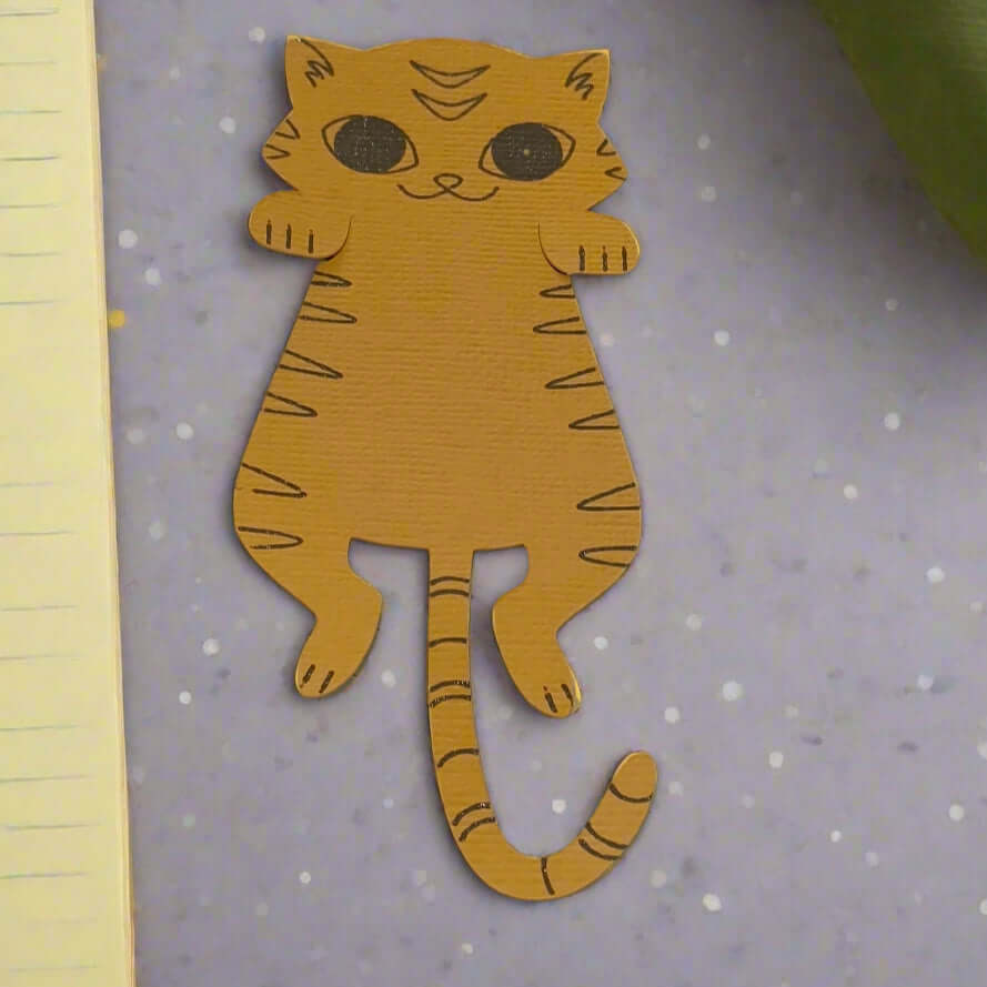 Top-down view of a cute orange tiger cat paper bookmark with black stripes, placed on a grey background beside a book and green leaves.