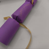 Handmade purple Diwali cracker tied with gold ribbon, foiled with “Happy Diwali” and diya motif, displayed against plain background.