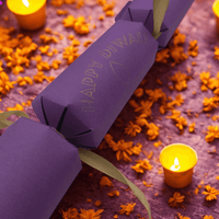 Handmade purple Diwali cracker with gold ribbon, surrounded by glowing tealight candles and marigold petals on a festive background.