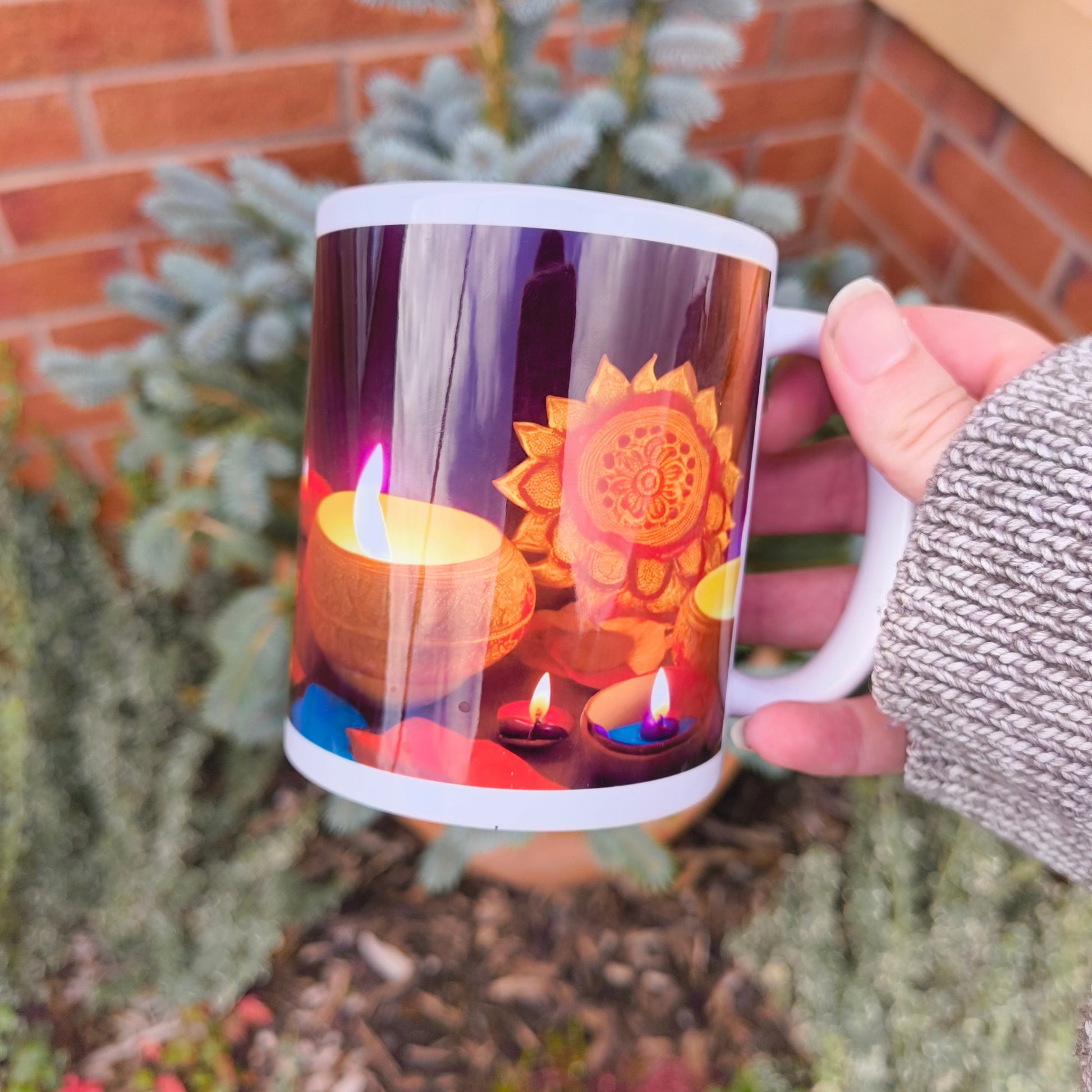 Hand holding a white Diwali-themed mug showing colourful diyas and a glowing mandala lantern design, photographed in a garden setting.