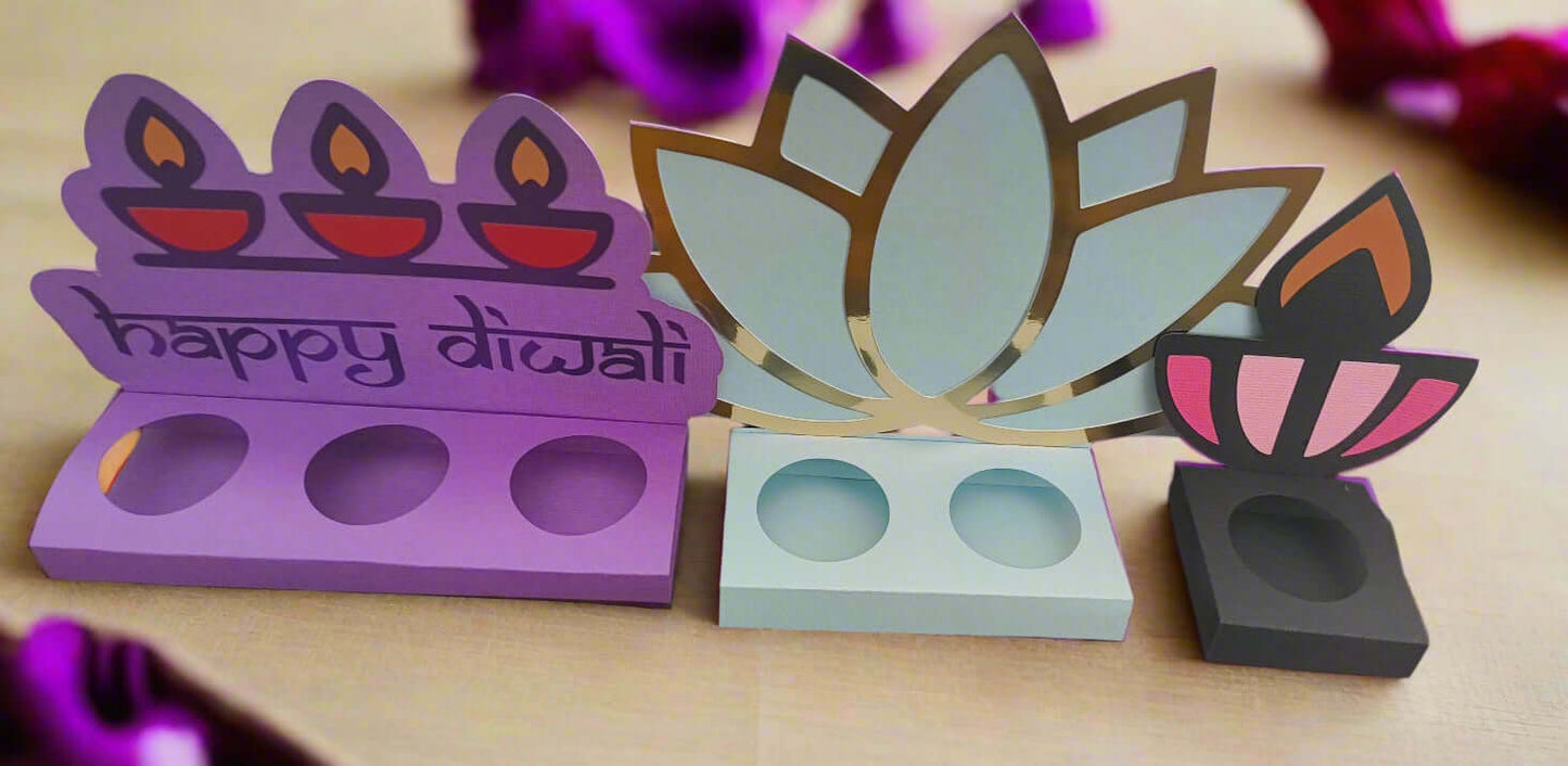 Front-facing image of a colourful set of three handcrafted Diwali sweet holders made from layered cardstock, featuring diya, lotus, and flame motifs on a warm-toned festive background.