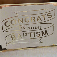 Elegant handmade baptism card with metallic gold foil text reading 'Congrats on Your Baptism', cut into textured white cardstock and displayed on a light wood surface.