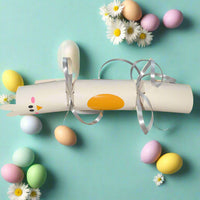 Handmade Easter cracker shaped like a chick, with cream cardstock, orange oval detail, and silver curling ribbon. Shown with pastel eggs and daisies for scale.